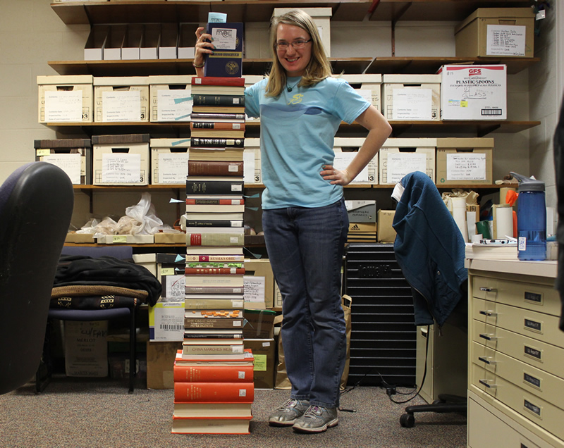 Student standing beside a stack of books as tall as she is.