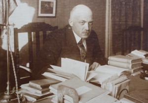 Black and white photo of balding man at a table surrounded by books.