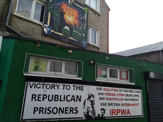 Buildings painted with murals that say "Join the IRA" and "Victory to the Republican Prisoners: End Isolation of Republicans, End Forced Strip Searching, End Controlled Movement, End British Internment IRPWA"