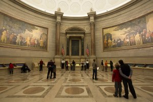 Rotunda for the Charters of Freedom in the National Archive, with murals on the wall illustrating the signing of the Declaration of Independence and the Constitution, with founding documents on display and visitors examining the display cases.